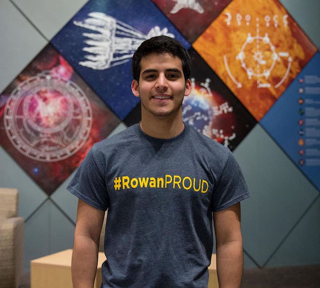Kevin wears a #RowanPROUD t-shirt in front of brightly colored background outside the planetarium