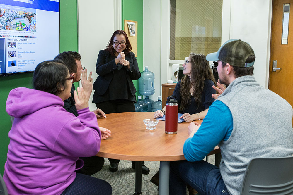 Small group of sign language students sitting at a conference table while professor stands and signs with them.