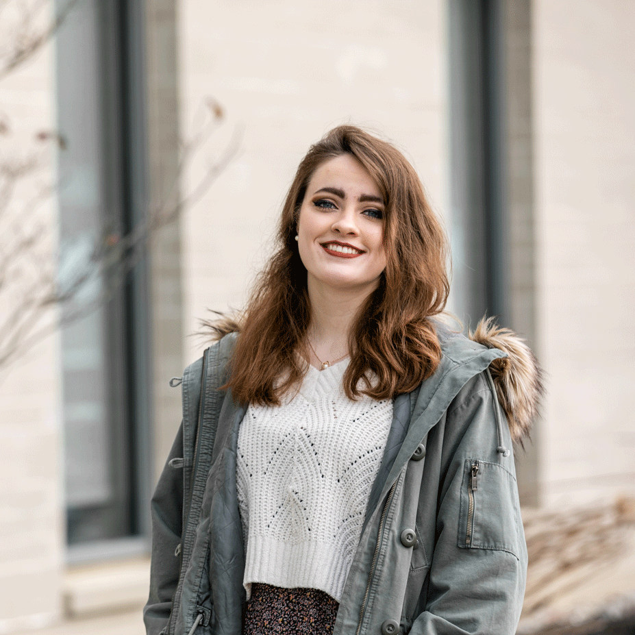Laura smiles wearing a coat outside an academic building on campus.