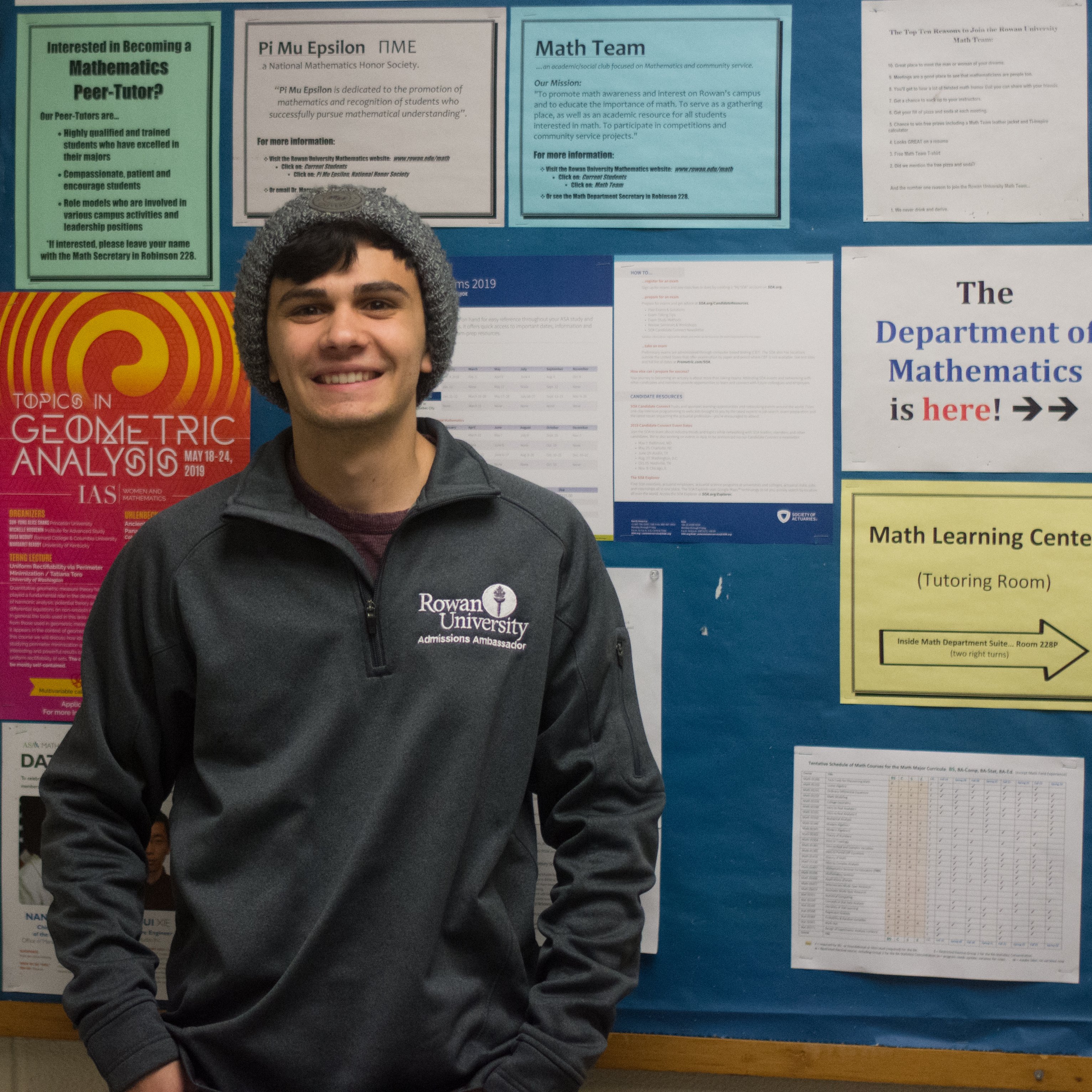 Jake stands in front of a math bulletin board, wearing a beanie on his head and a gray pullover, hands in his pockets. 