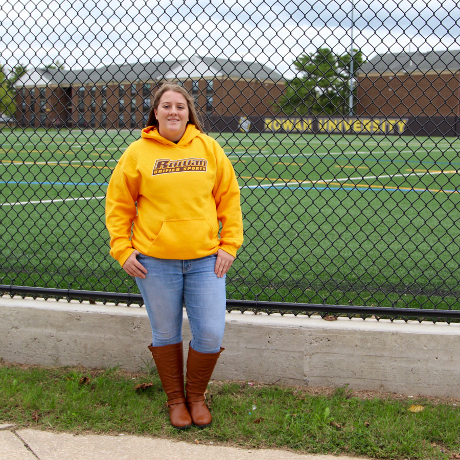 Maribeth stands outside of a brick dorm at Rowan University with her left hand on her hip, wearing a gray #RowanPROUD t-shirt.