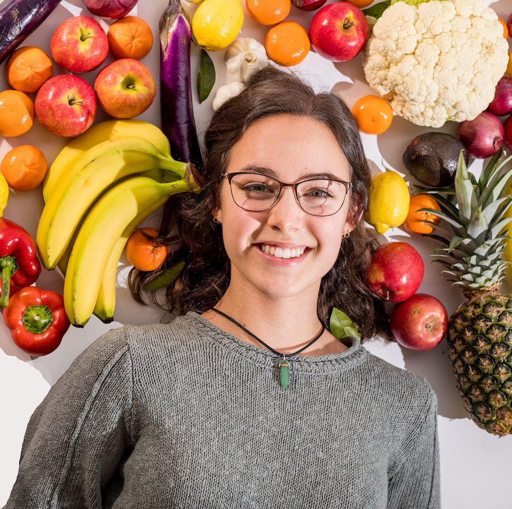 Hannah smiles with a halo of multi-colored fruits and vegetables around her head.
