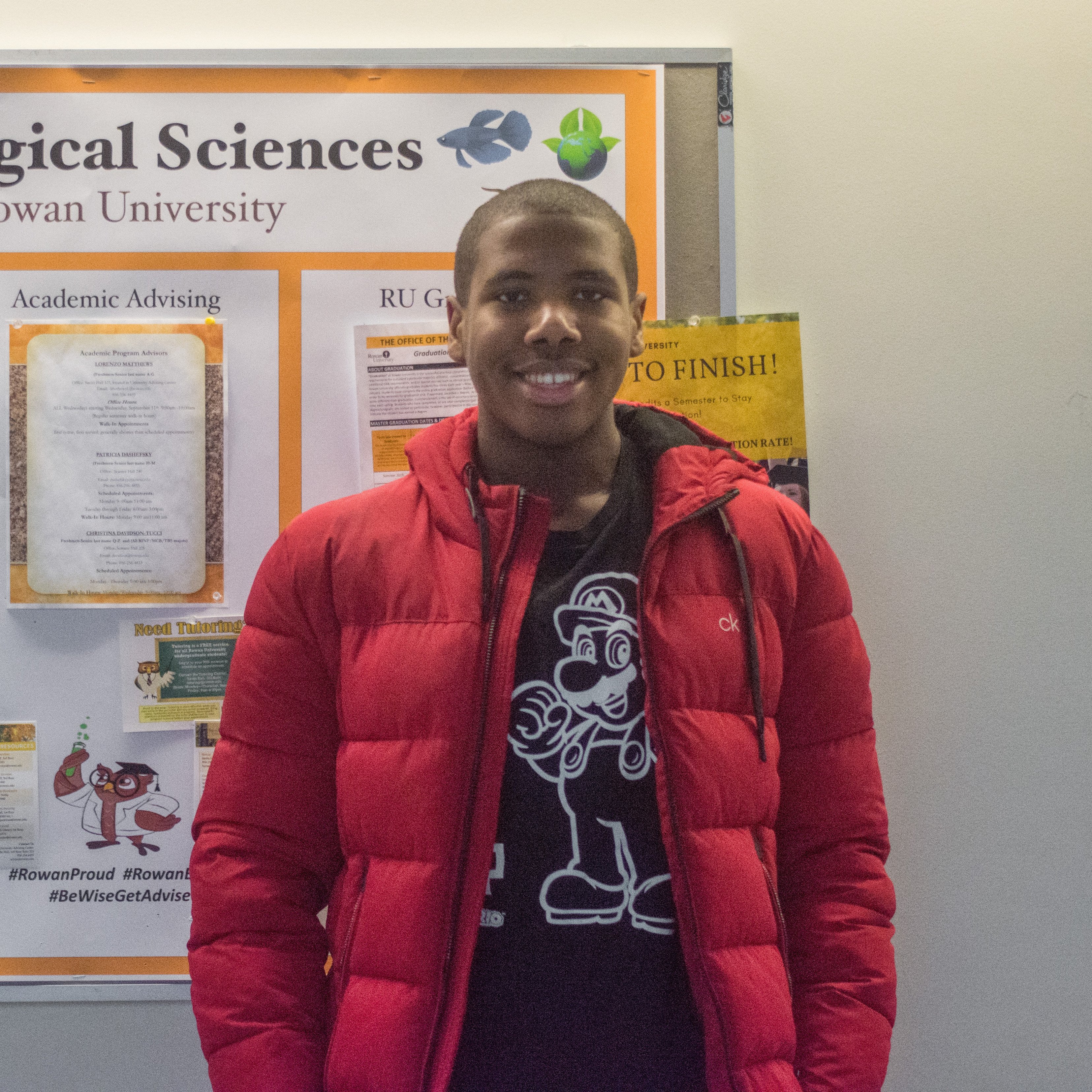 Sean wears a red puffy coat in front of a science bulletin board. 