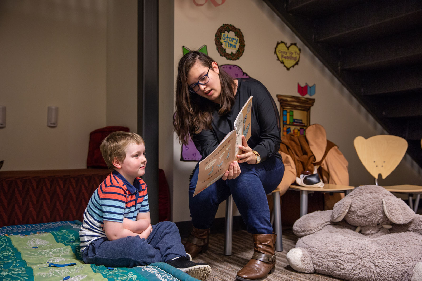 college student sitting on chair reading to small child sitting on rug