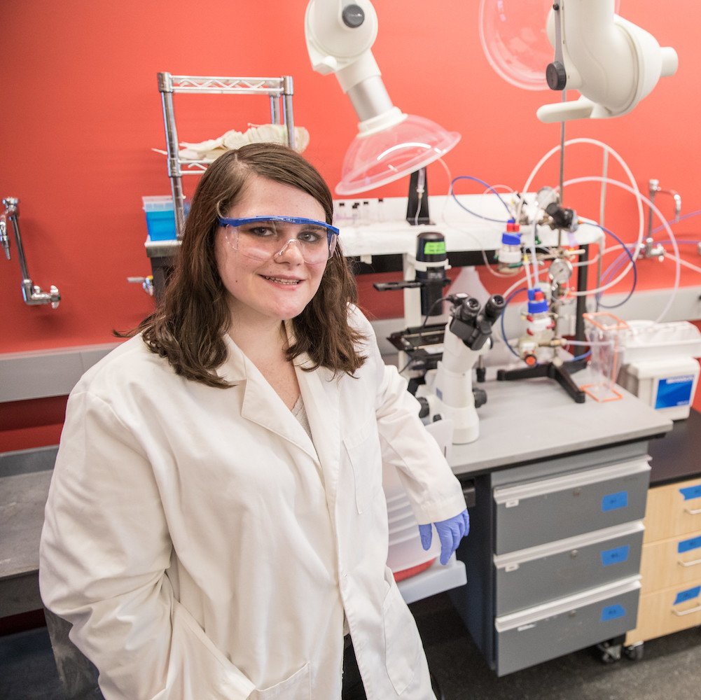Marissa stands next to equipment, elbow resting on it, while wearing goggles, lab coat and gloves.
