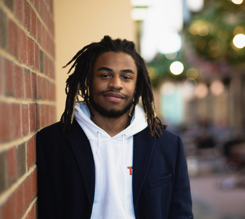 Rowan University student Cristofer smiles at the camera posing for a portrait against a brick wall.