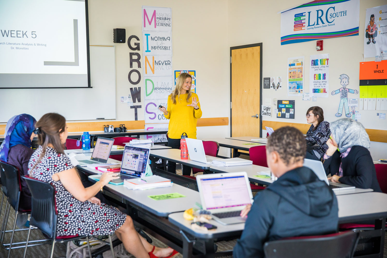 educator stands in brightly decorated classroom leading discussion amongst PhD students.