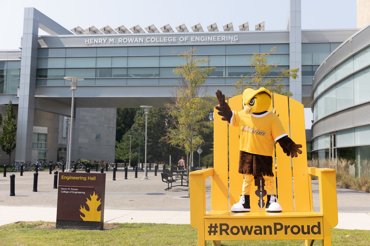 Rowan University owl mascot Whoo RU stands on an oversized yellow chair that says #RowanPROUD, while in front of the engineering building. 