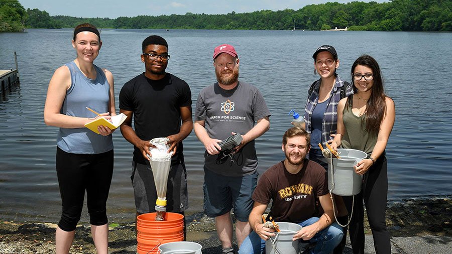 Biological science majors stand with a professor in front of a lake for an out of class experience.
